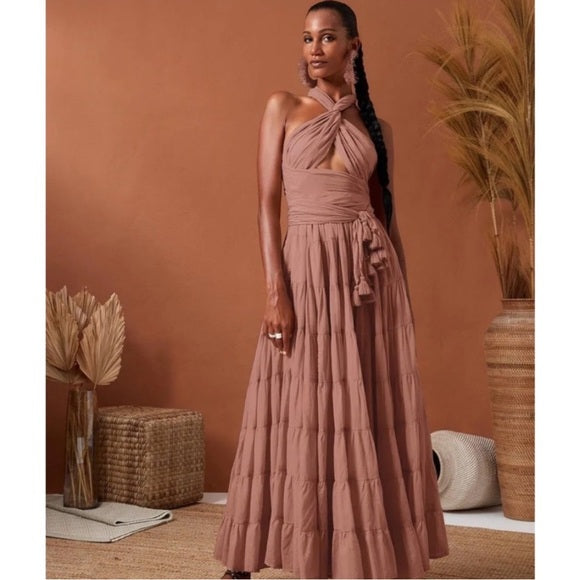 She's wearing a flowing, dusty rose or terracotta-colored maxi dress with a crisscross halter-style top and a high-waisted, belted skirt that has a tiered, ruffled design.