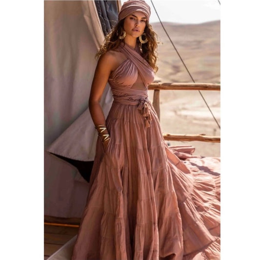 She's wearing a flowing, dusty rose or terracotta-colored maxi dress with a crisscross halter-style top and a high-waisted, belted skirt that has a tiered, ruffled design.